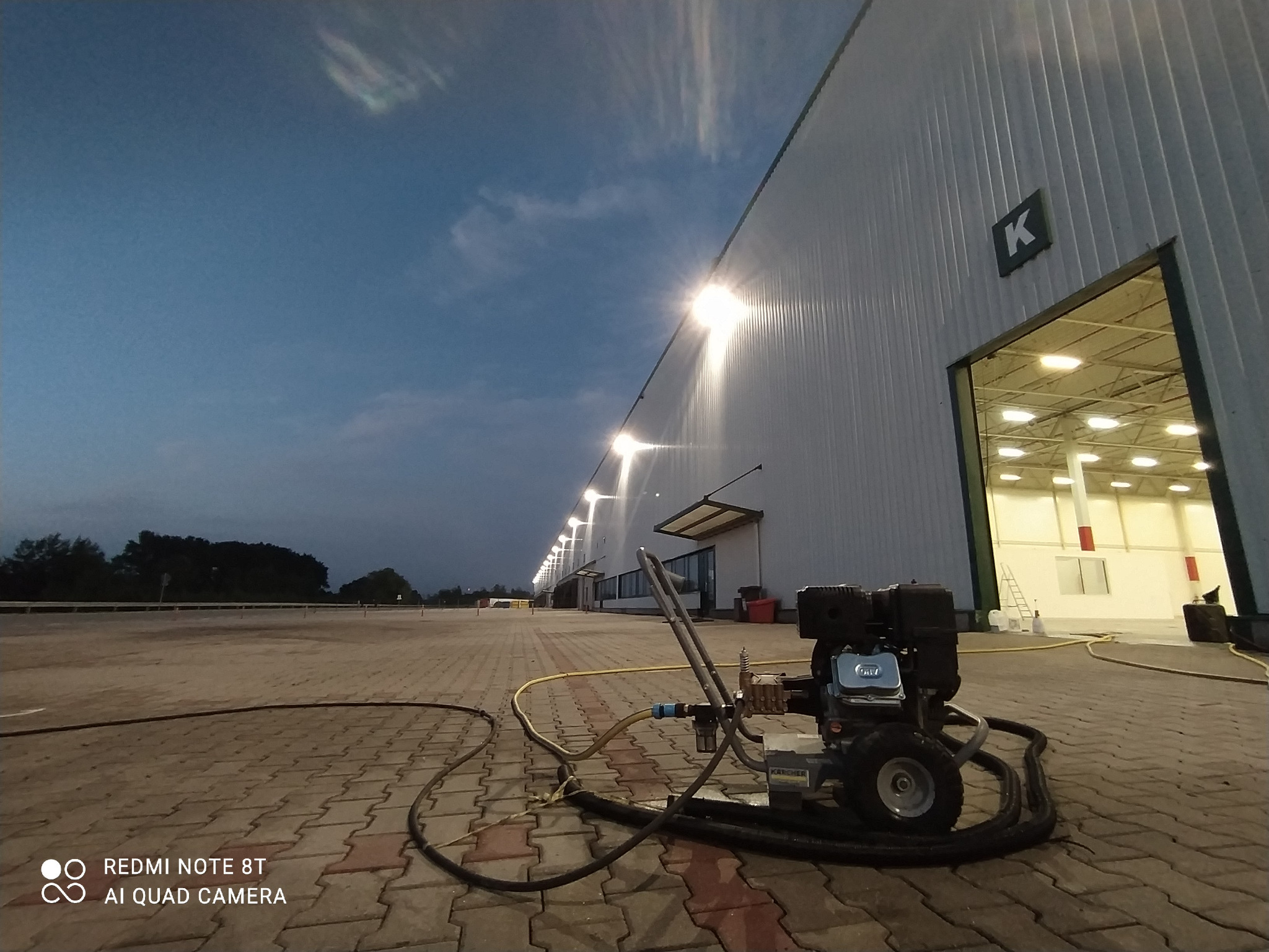 Profesjonalna myjka ciśnieniowa Karcher na kostce brukowej przed halą magazynową z otwartymi wrotami, widoczne oświetlenie zewnętrzne i fragment zachmurzonego nieba.