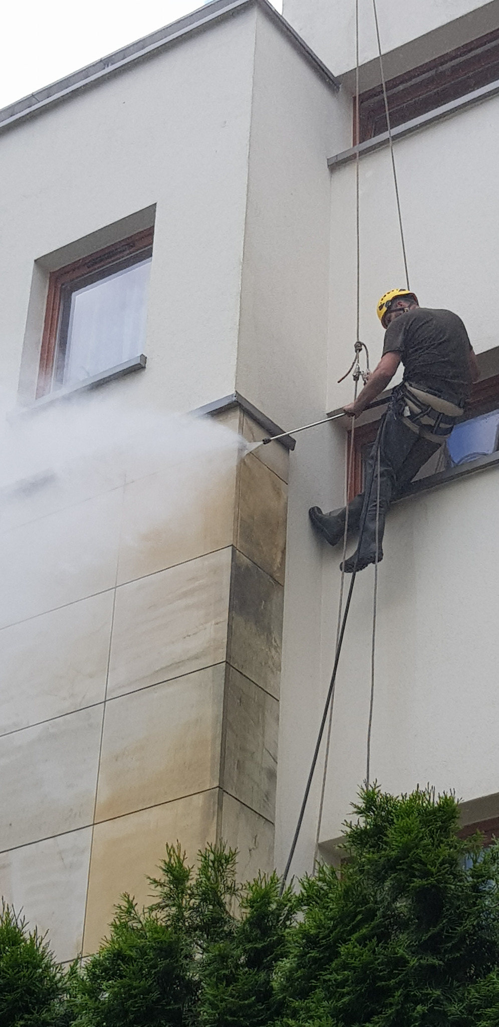 Pracownik w uprzęży, z żółtym kaskiem, czyści elewację budynku z wysokości za pomocą myjki ciśnieniowej, widoczny strumień wody i fragment elewacji przed i po czyszczeniu.