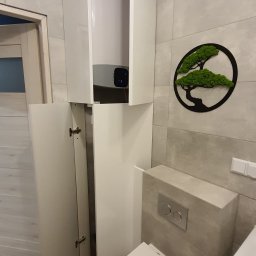 Lubomir Lubomir - Biała szafka łazienkowa na wymiar z otwartymi drzwiami, ukrywająca bojler, obok podwieszana toaleta i dekoracja ścienna z mchu.