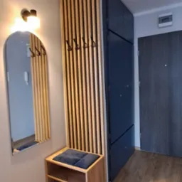 Nowoczesny przedpokój z szafą w kolorze granatowym, drewnianym panelem ściennym z wieszakami, lustrem w drewnianej ramie oraz siedziskiem z półkami na buty, oświetlonym kinkietem.