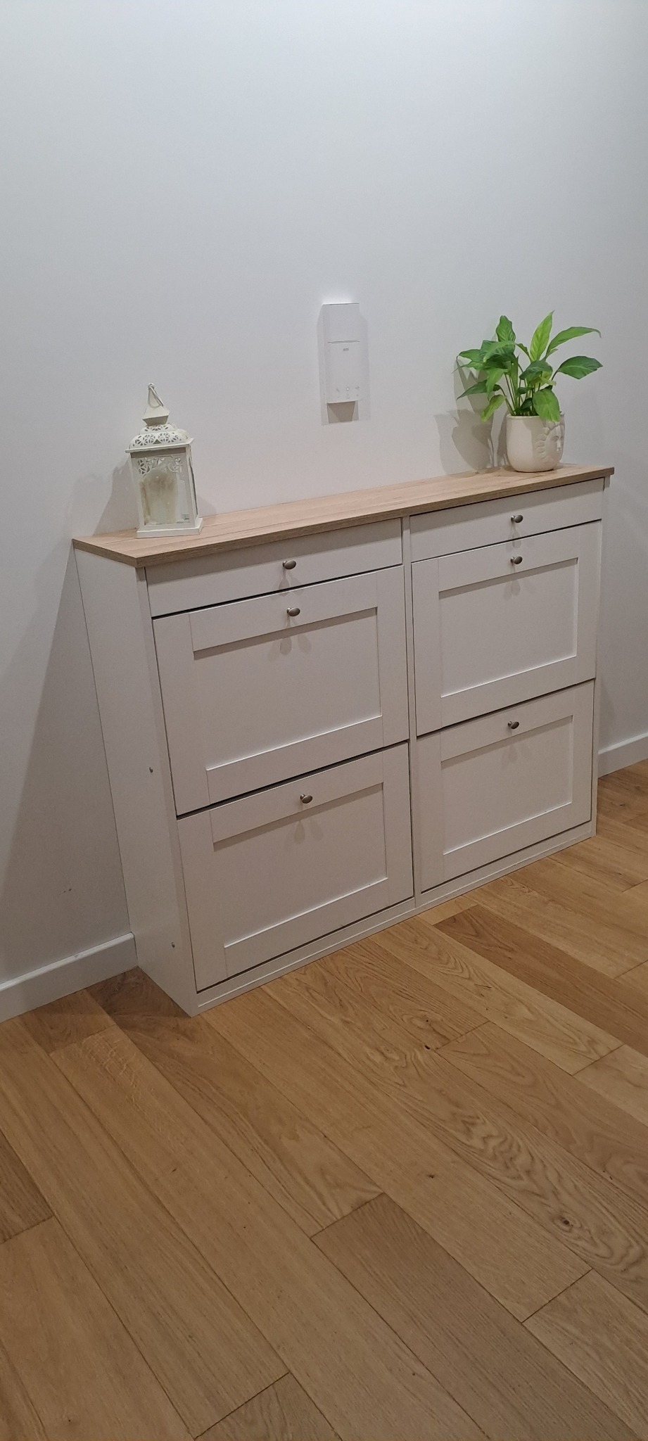 Dwu-segmentowa szafka na buty w kolorze jasnoszarym z drewnianym blatem, na blacie dekoracyjna latarenka i doniczka z zieloną rośliną, na tle biała ściana i parkiet.