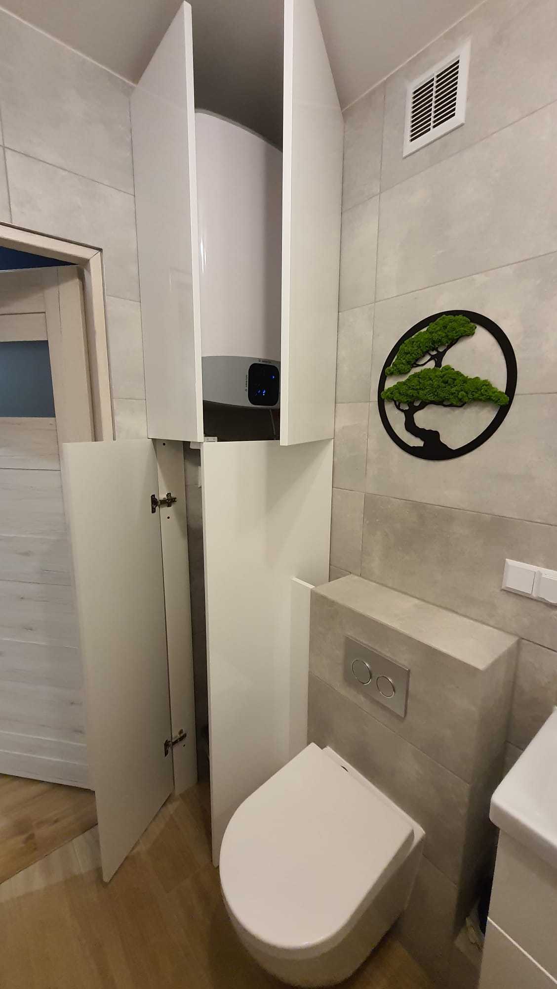 Biała szafka łazienkowa na wymiar z otwartymi drzwiami, ukrywająca bojler, obok podwieszana toaleta i dekoracja ścienna z mchu.