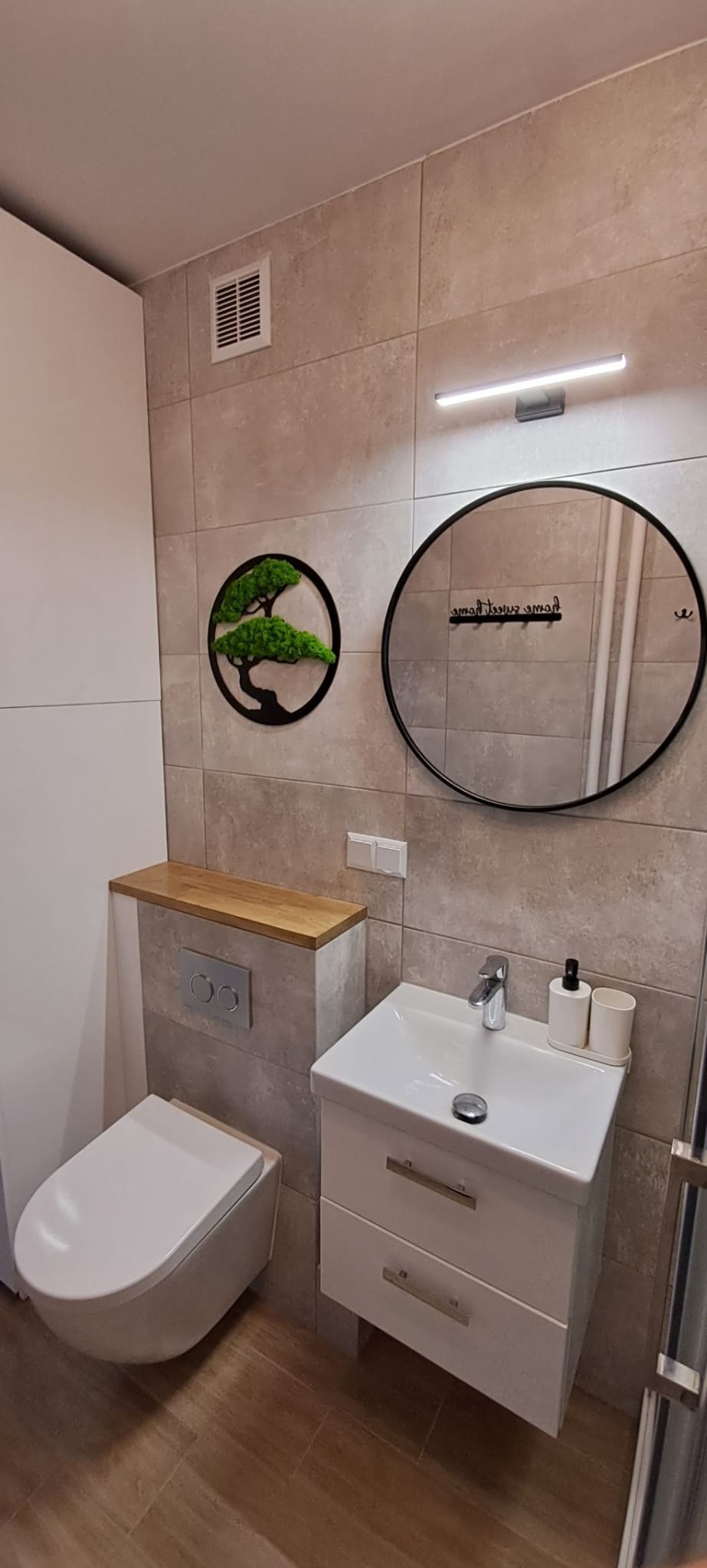 Nowoczesna łazienka z podwieszaną toaletą, umywalką z szafką, okrągłym lustrem z haczykami i dekoracją ścienną imitującą drzewko bonsai.