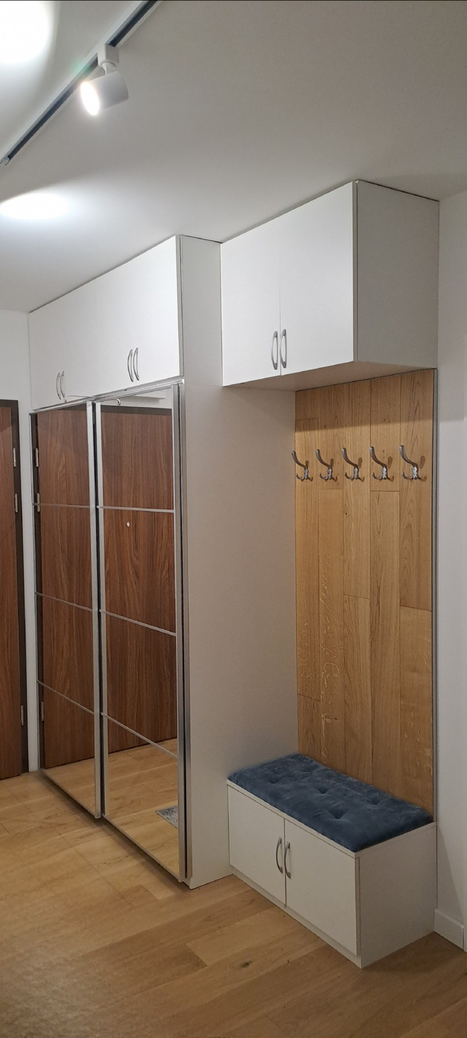 Wnękowa szafa z przesuwnymi lustrzanymi drzwiami, połączona z panelem w kolorze drewna z wieszakami i szafką z siedziskiem w przedpokoju.