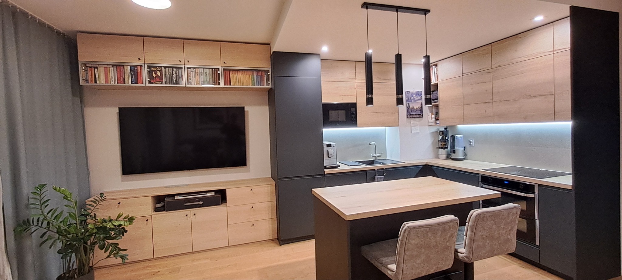 Nowoczesna kuchnia z wyspą, fronty w odcieniach drewna i szarości, oświetlenie LED podszafkowe, regał z książkami nad telewizorem, dwie krzesła barowe z tapicerką.