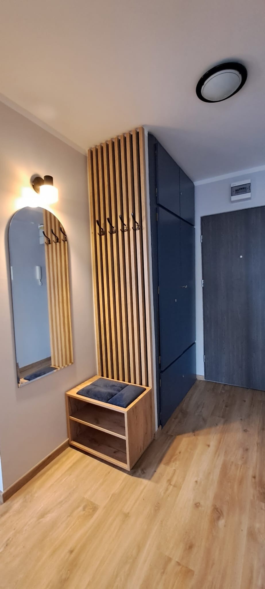 Nowoczesny przedpokój z szafą w kolorze granatowym, drewnianym panelem ściennym z wieszakami, lustrem w drewnianej ramie oraz siedziskiem z półkami na buty, oświetlonym kinkietem.