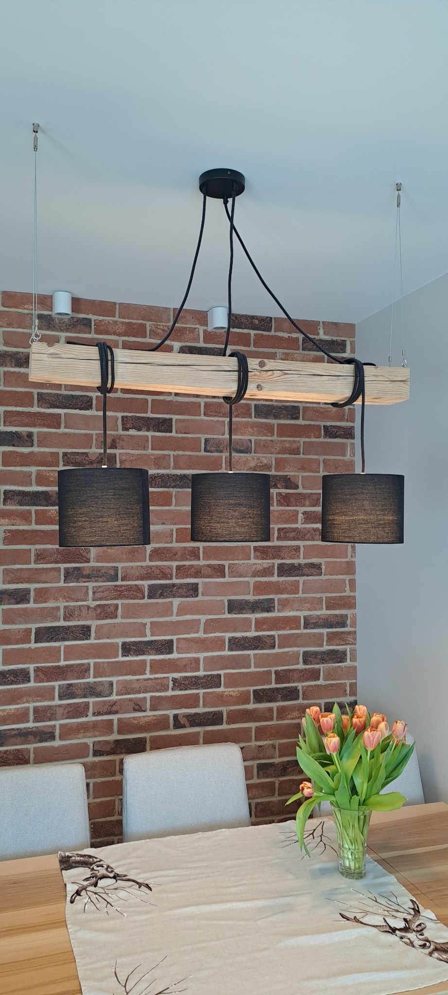 Drewniana lampa wisząca z trzema czarnymi abażurami, zamontowana nad stołem z dekoracyjnym bieżnikiem i bukietem tulipanów na tle ściany z cegły.