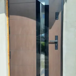 Nowoczesne brązowe drzwi wejściowe z pionowym przeszkleniem, widoczne pianka montażowa wokół ościeżnicy, płytki na podłodze.