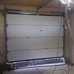 LS Serwis Montaż Bramy Okna Drzwi Sławomir Lewandowski - Zamknięta, biała brama garażowa segmentowa z widocznymi elementami konstrukcyjnymi i oświetleniem roboczym w surowym garażu.