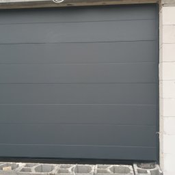 LS Serwis Montaż Bramy Okna Drzwi Sławomir Lewandowski - Zamknięta, segmentowa brama garażowa w kolorze antracytowym, widoczna w surowym otoczeniu budowy, z białymi bloczkami betonowymi i drewnianymi paletami w tle.