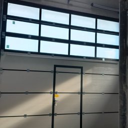 LS Serwis Montaż Bramy Okna Drzwi Sławomir Lewandowski - Zamknięta brama segmentowa garażowa z oknami i drzwiami wejściowymi, widok od wewnątrz, surowy betonowy sufit, fragment rusztowania po prawej stronie.