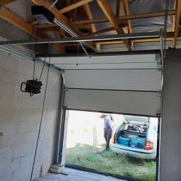 LS Serwis Montaż Bramy Okna Drzwi Sławomir Lewandowski - Montaż segmentowej bramy garażowej: widoczny mechanizm otwierania pod sufitem, częściowo otwarta brama z widokiem na samochód z narzędziami w bagażniku, surowy stan garażu.