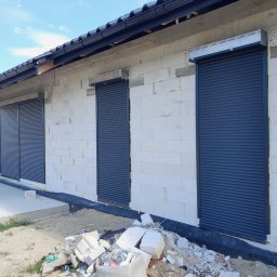 LS Serwis Montaż Bramy Okna Drzwi Sławomir Lewandowski - Nowo wybudowany dom z trzema oknami zabezpieczonymi szarymi roletami zewnętrznymi, widoczne materiały budowlane i samochód w tle.