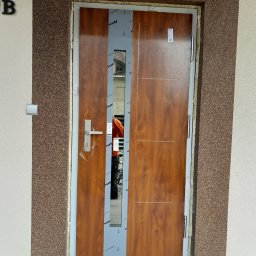 LS Serwis Montaż Bramy Okna Drzwi Sławomir Lewandowski - Brązowe drzwi wejściowe z drewnopodobnym wykończeniem, zabezpieczone folią ochronną, zamontowane w elewacji budynku z dekoracyjnym tynkiem.