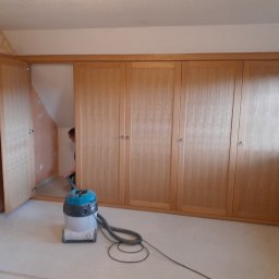 LS Serwis Montaż Bramy Okna Drzwi Sławomir Lewandowski - Drewniana zabudowa szafy wnękowej z otwartymi drzwiami po lewej stronie, widoczny odkurzacz i osoba sprzątająca wewnątrz szafy.