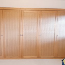 LS Serwis Montaż Bramy Okna Drzwi Sławomir Lewandowski - Jasnobrązowa, drewniana szafa wnękowa z sześciorgiem drzwi, z drobnymi, okrągłymi uchwytami, częściowo zasłonięta przez osobę w roboczym ubraniu.