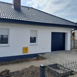 Nowoczesny dom jednorodzinny w Henrykowie z czarną bramą garażową, oknami z roletami zewnętrznymi i czarnym dachem z dachówki ceramicznej, widok z zewnątrz.