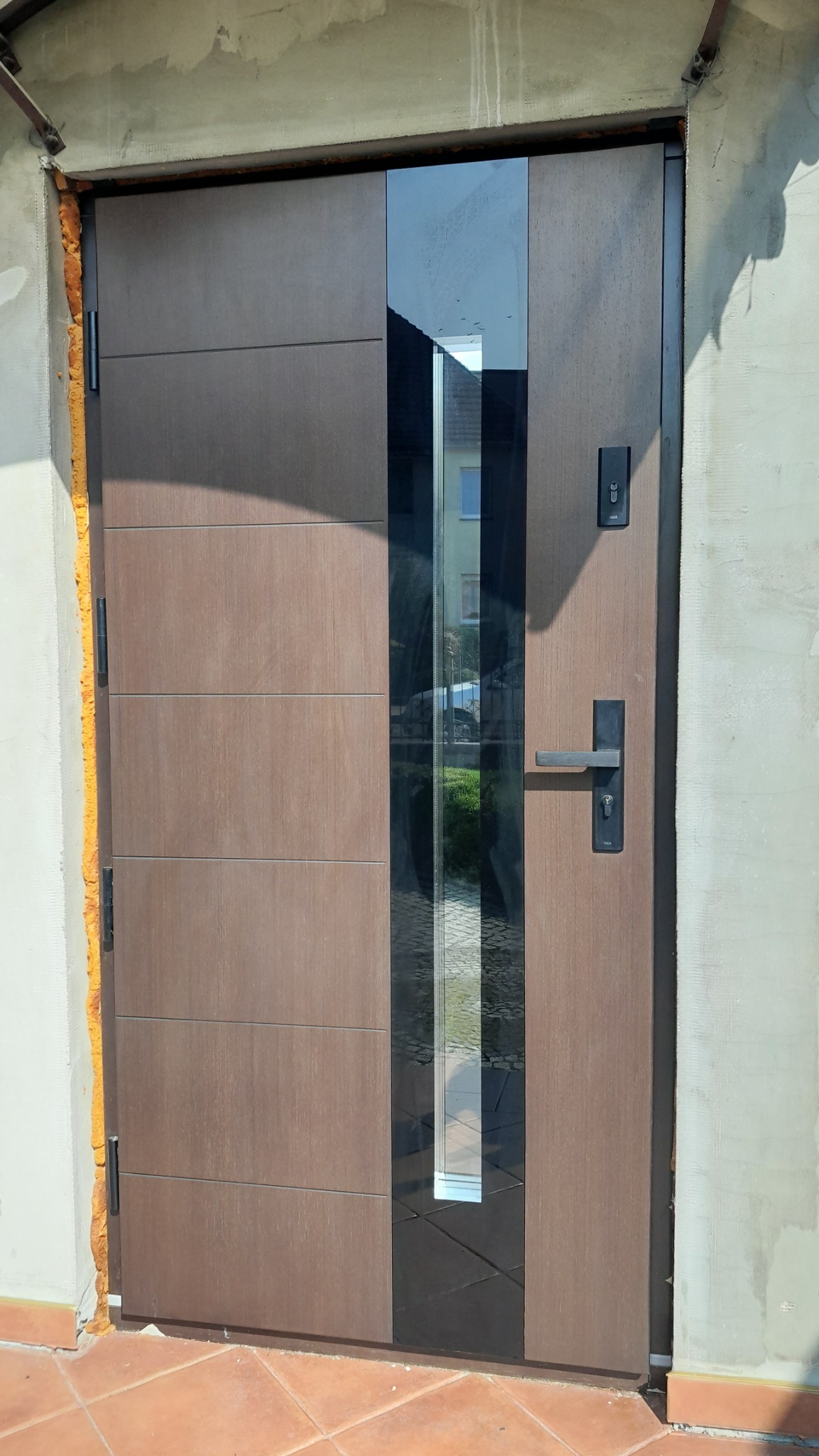 Nowoczesne brązowe drzwi wejściowe z pionowym przeszkleniem, widoczne pianka montażowa wokół ościeżnicy, płytki na podłodze.