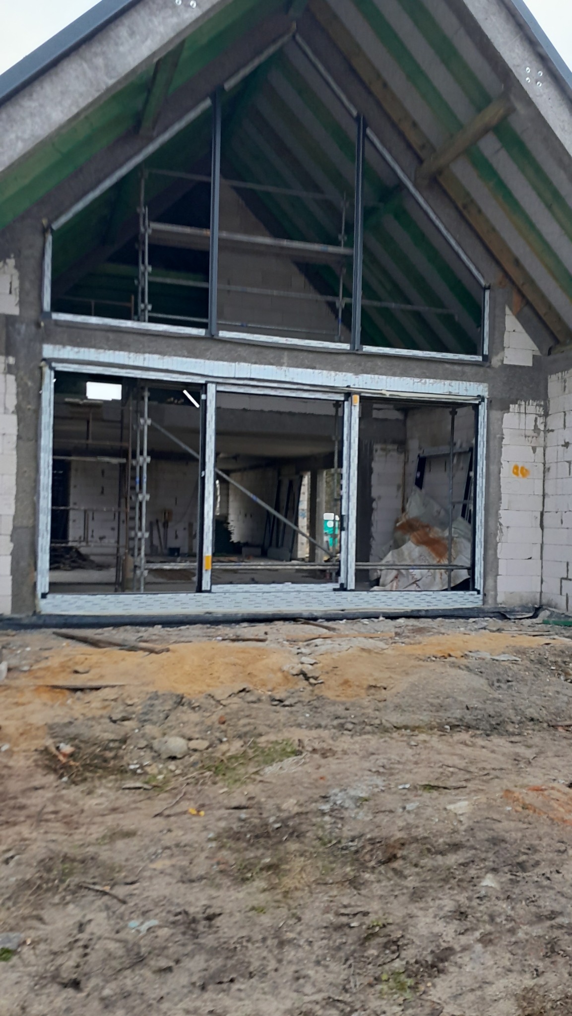 Surowy stan budynku z zamontowanymi oknami PCV i ramami drzwi przesuwnych, widoczne rusztowania wewnątrz i niezagospodarowany teren przed budynkiem.