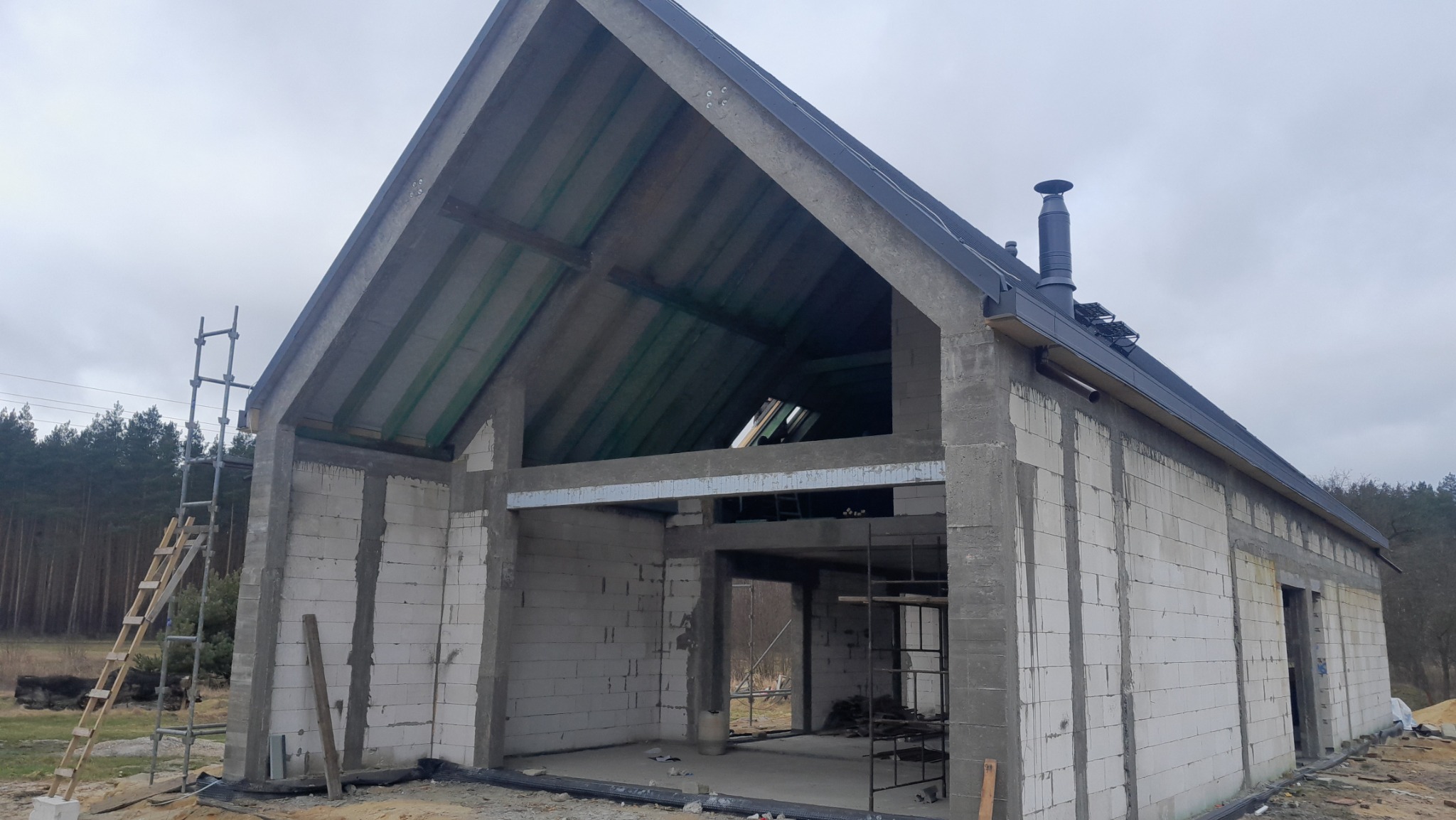 Budowa domu jednorodzinnego w stanie surowym otwartym, widoczne ściany z betonu komórkowego, drewniana konstrukcja dachu z oknem połaciowym, komin i rusztowania.