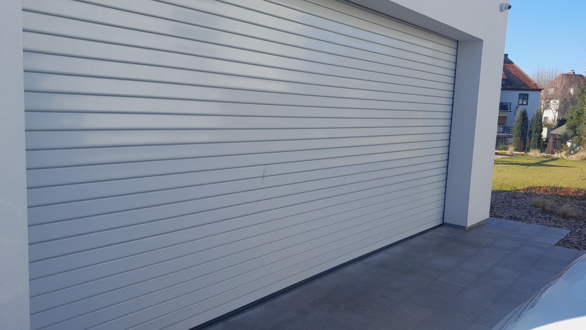 Zamknięta, pozioma, segmentowa brama garażowa w kolorze jasnoszarym, z delikatnym zarysowaniem na powierzchni, widoczna z zewnątrz budynku w słoneczny dzień.