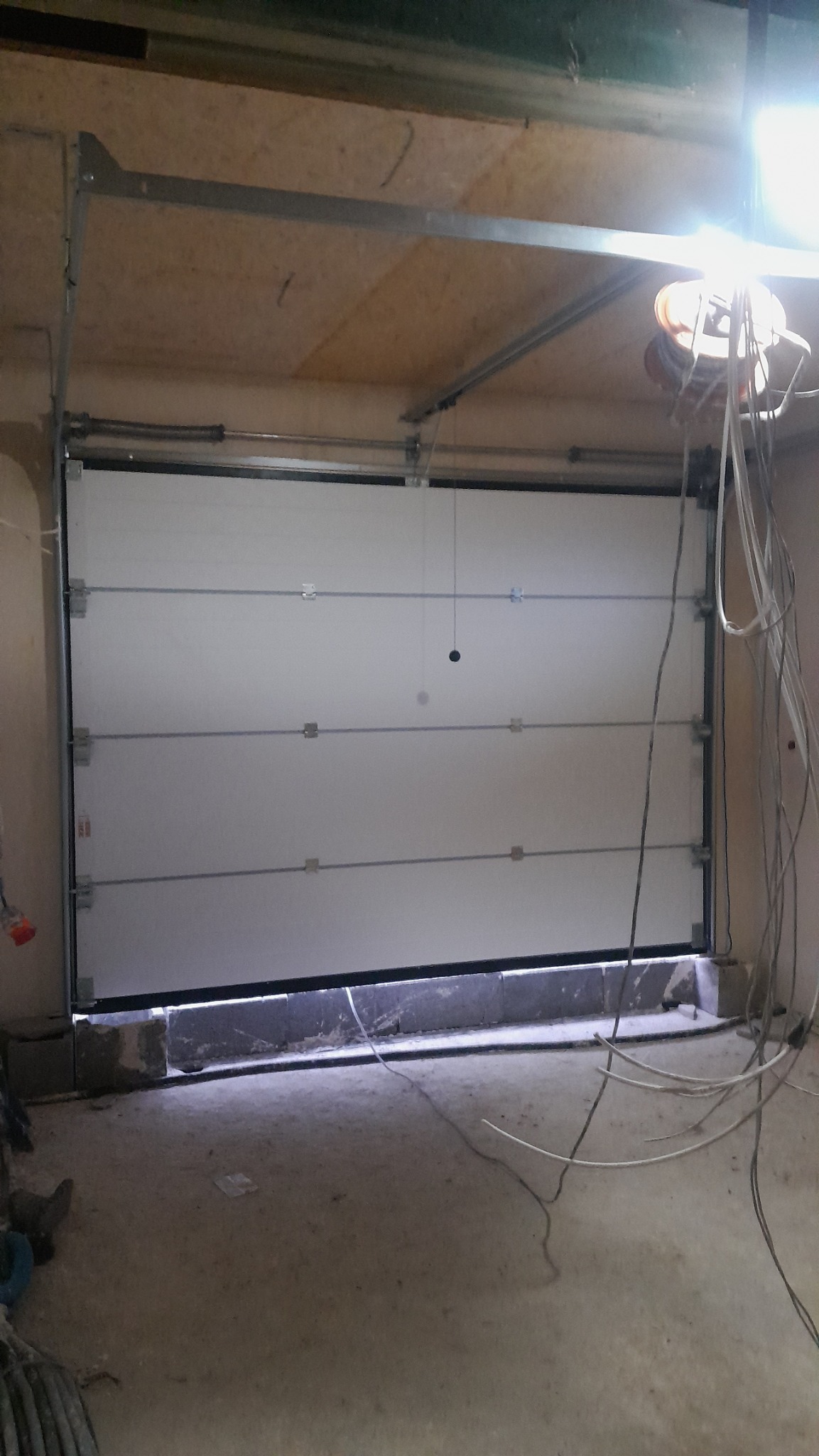 Zamknięta, biała brama garażowa segmentowa z widocznymi elementami konstrukcyjnymi i oświetleniem roboczym w surowym garażu.