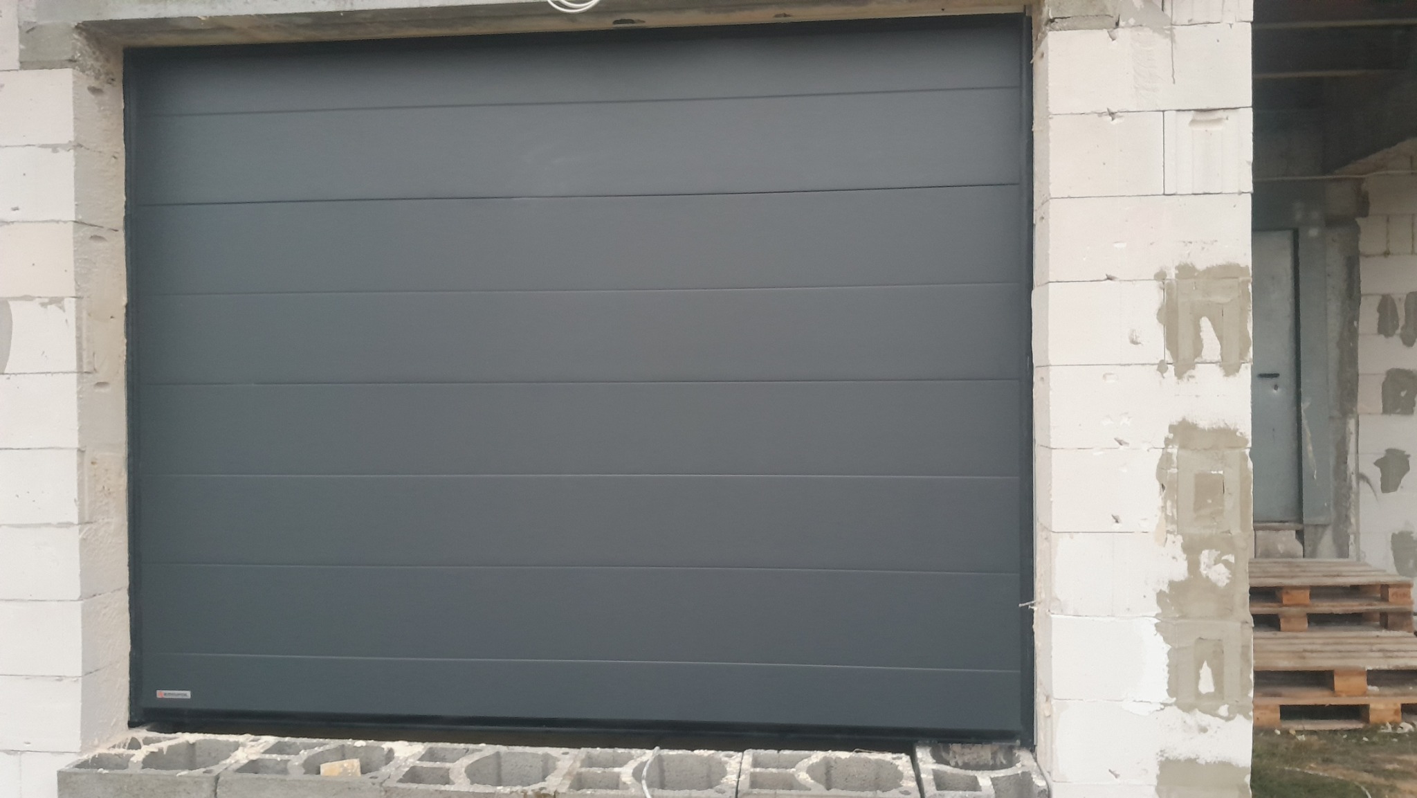Zamknięta, segmentowa brama garażowa w kolorze antracytowym, widoczna w surowym otoczeniu budowy, z białymi bloczkami betonowymi i drewnianymi paletami w tle.
