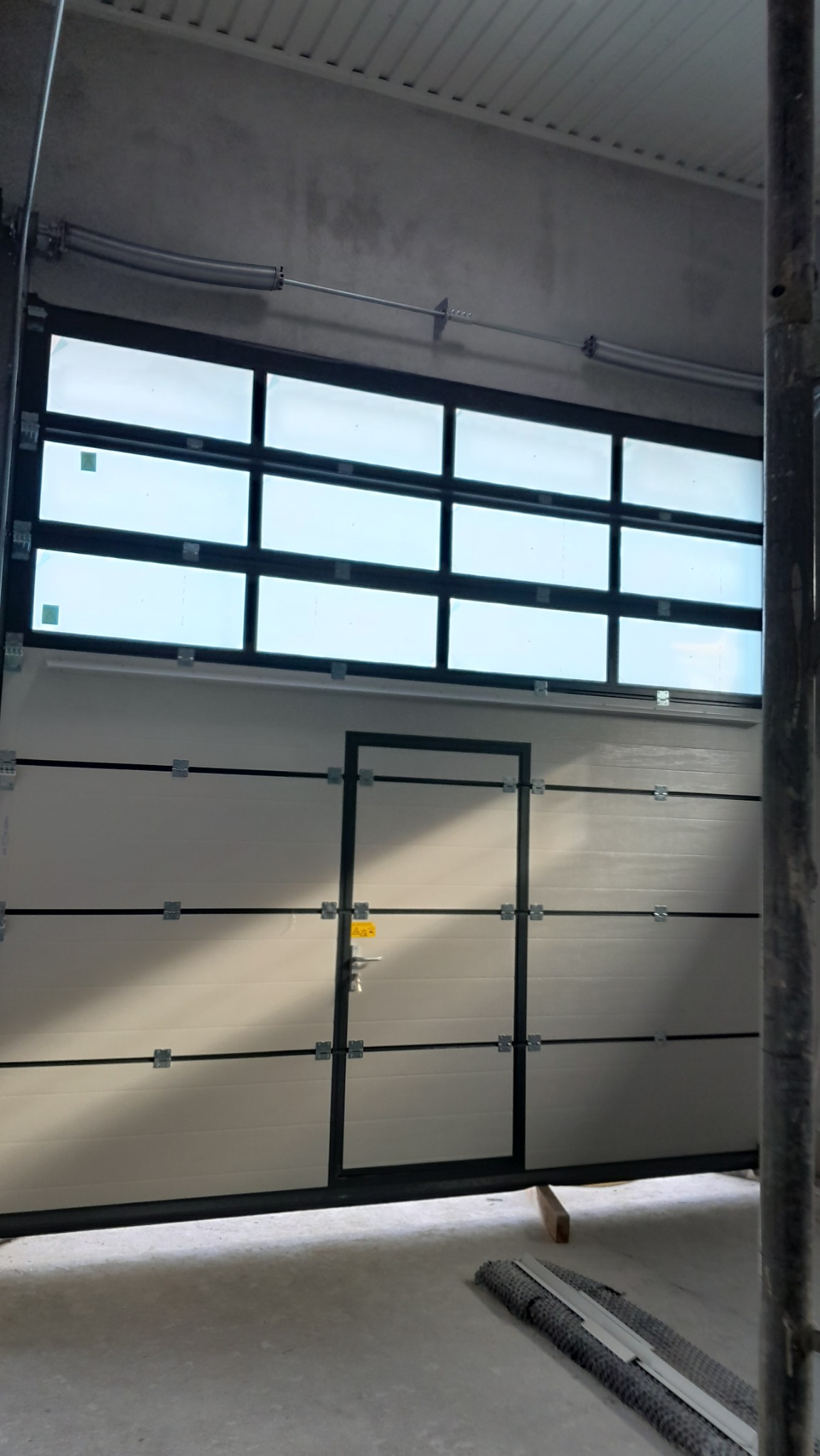 Zamknięta brama segmentowa garażowa z oknami i drzwiami wejściowymi, widok od wewnątrz, surowy betonowy sufit, fragment rusztowania po prawej stronie.