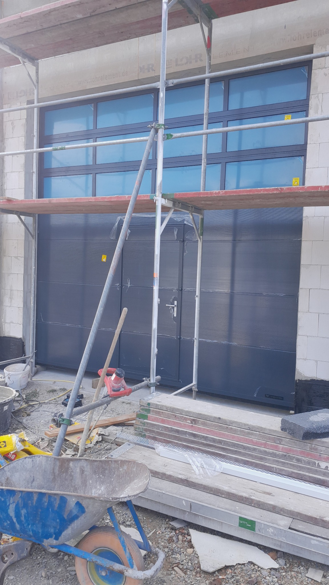 Szara segmentowa brama garażowa z oknami u góry, widoczna z rusztowania na placu budowy, z taczką i narzędziami w pierwszym planie.