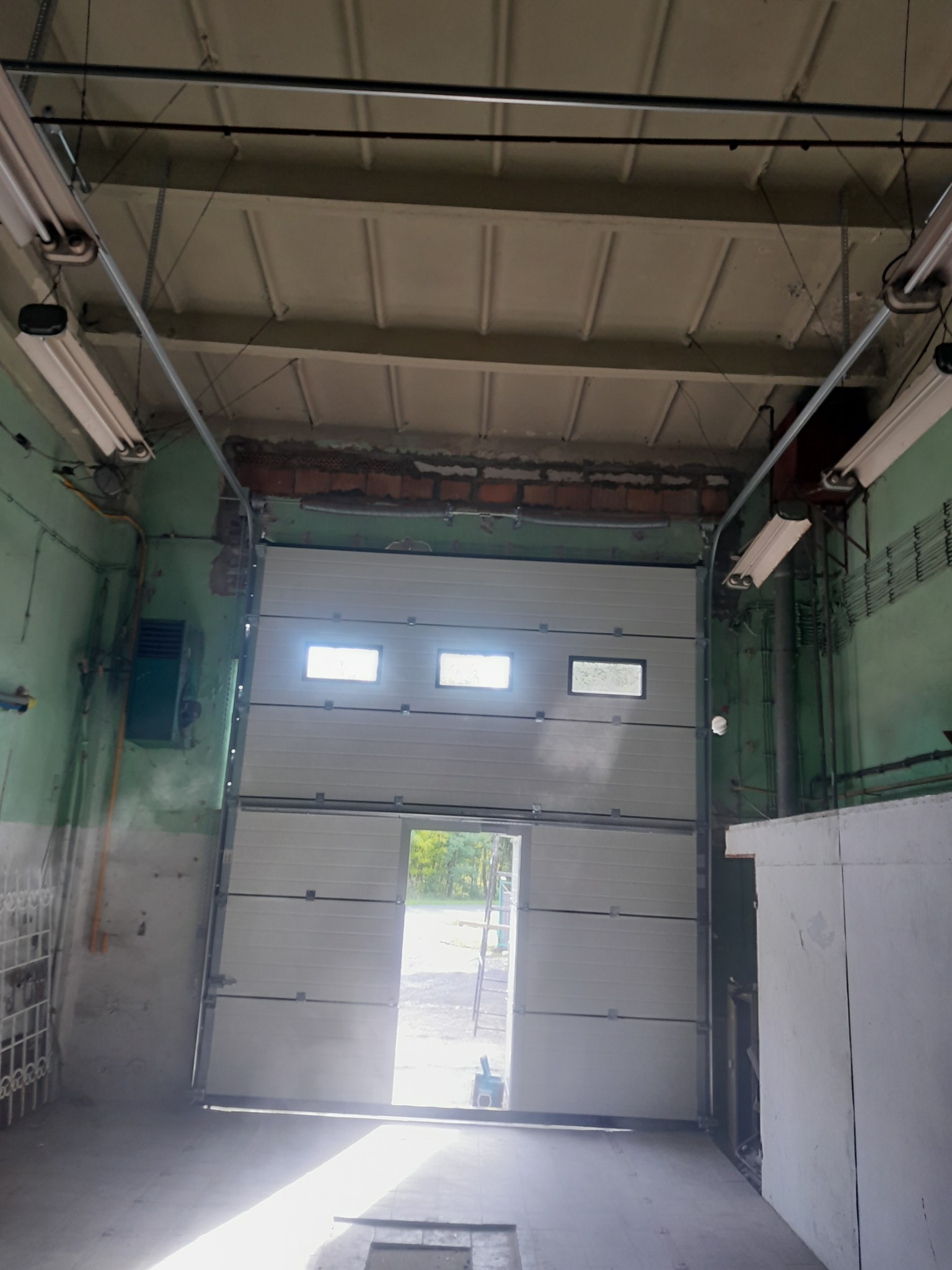 Otwarta segmentowa brama garażowa z trzema małymi oknami, widok z wnętrza garażu o surowym wykończeniu, z widocznym fragmentem podwórka przez otwór bramy.