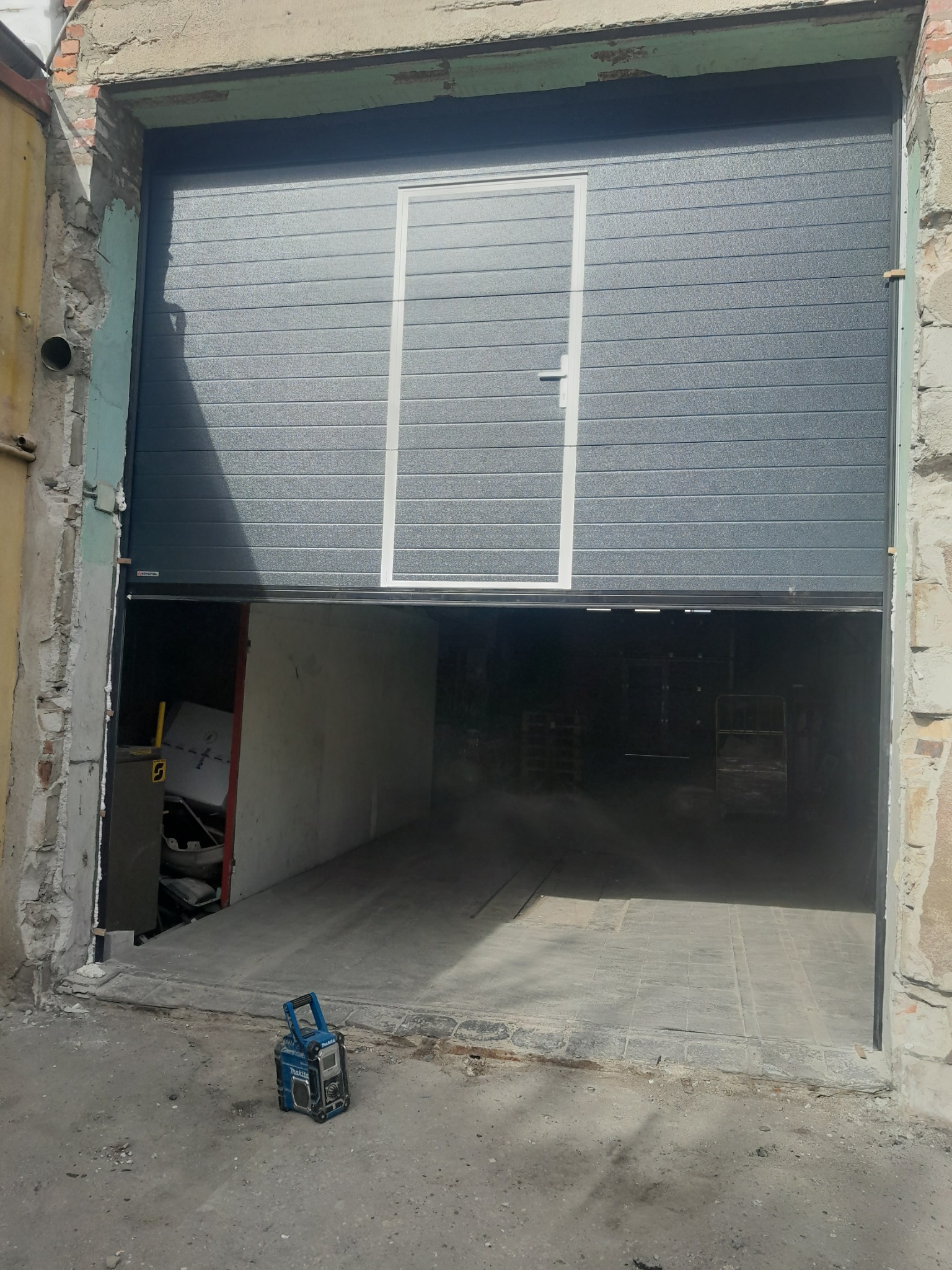Szara segmentowa brama garażowa z wbudowanymi drzwiami, częściowo otwarta, z widocznym wnętrzem garażu i radiem budowlanym Makita na posadzce przed wjazdem.