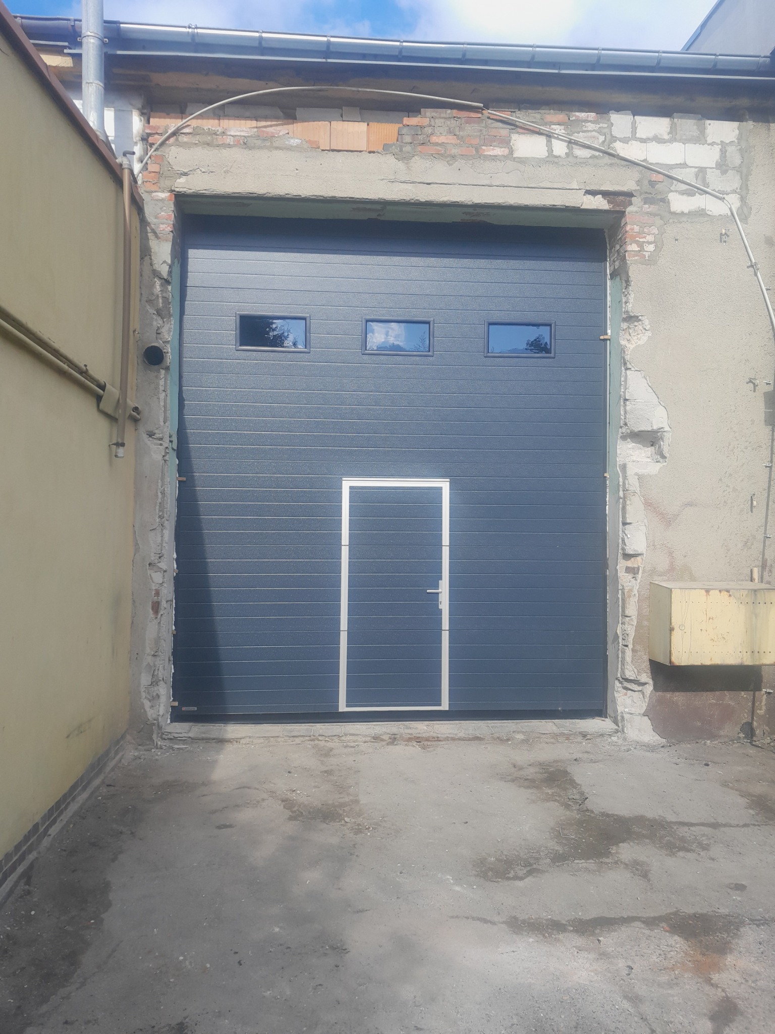 Grafitowa brama garażowa segmentowa z trzema małymi oknami i drzwiami wejściowymi, zamontowana w surowym otoczeniu z widocznymi uszkodzeniami muru.
