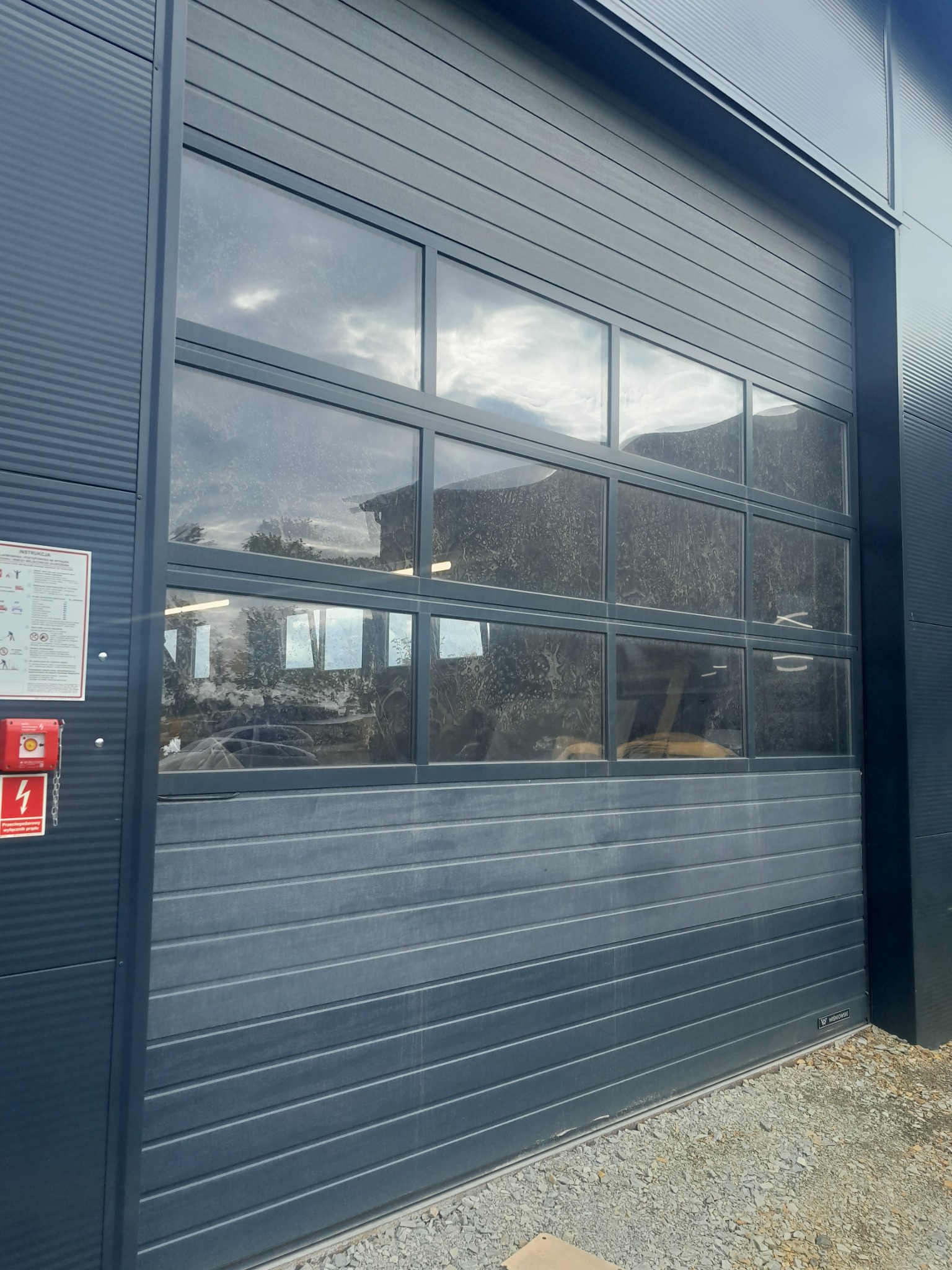 Szara segmentowa brama garażowa z oknami, widoczne zabrudzenia na szybach, obok tablica informacyjna i przycisk alarmowy.