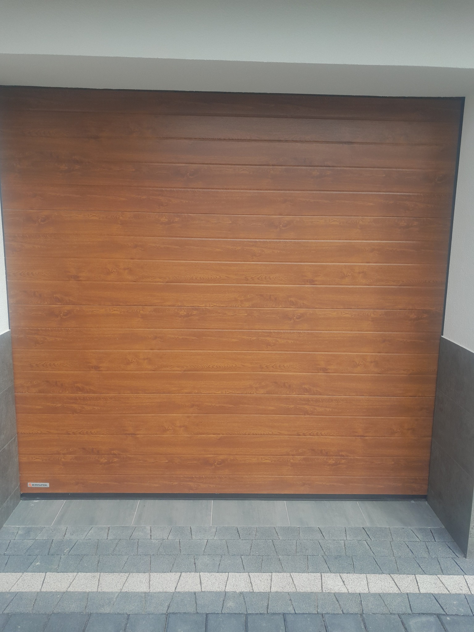 Zamknięta brama garażowa segmentowa o strukturze drewna, widoczna z zewnątrz, z szarą kostką brukową przed nią i fragmentem ściany po bokach.