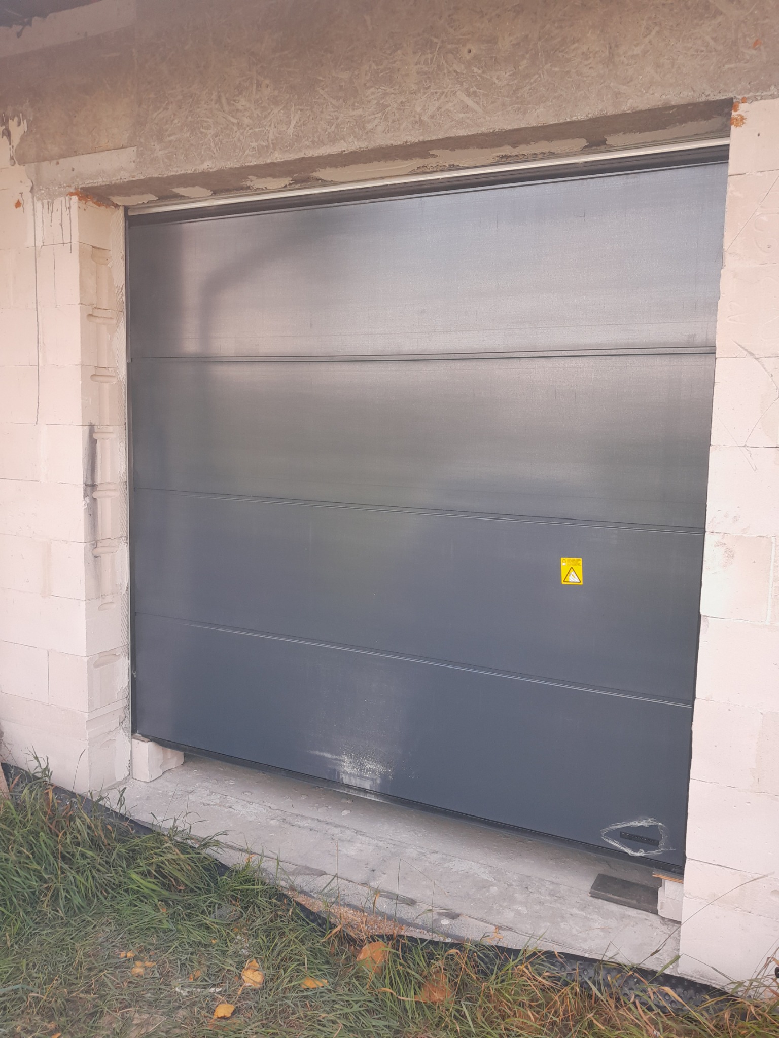 Zamknięta, szara brama garażowa segmentowa z żółtą naklejką ostrzegawczą, w surowym otworze budynku z białych bloczków, widoczne ślady montażu i trawa u dołu.