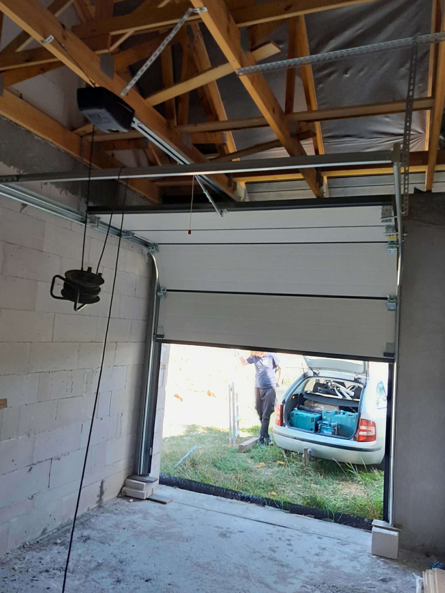 Montaż segmentowej bramy garażowej: widoczny mechanizm otwierania pod sufitem, częściowo otwarta brama z widokiem na samochód z narzędziami w bagażniku, surowy stan garażu.