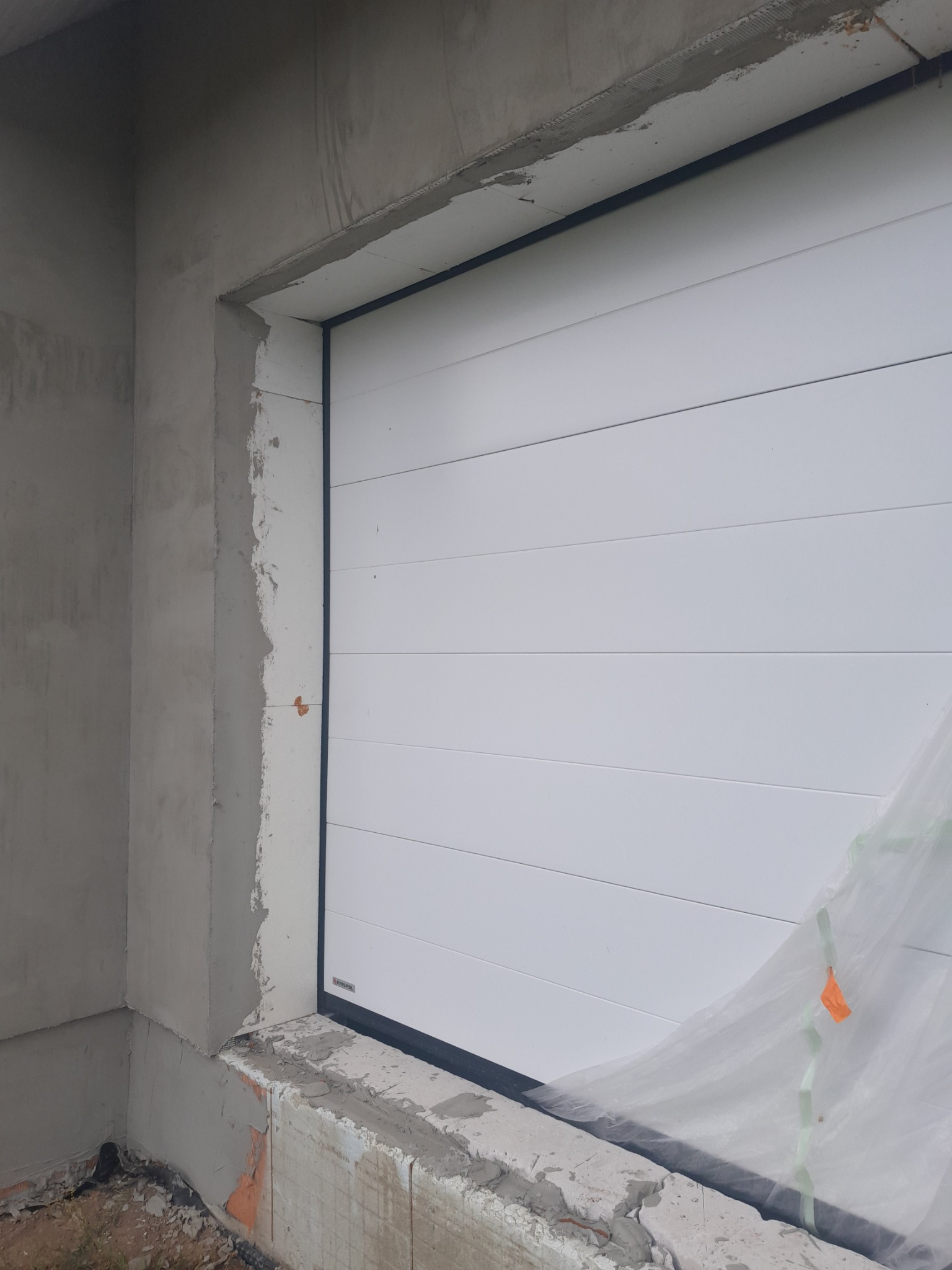 Biała segmentowa brama garażowa z czarną ramą, w trakcie montażu w surowym otoczeniu, zabezpieczona folią ochronną.