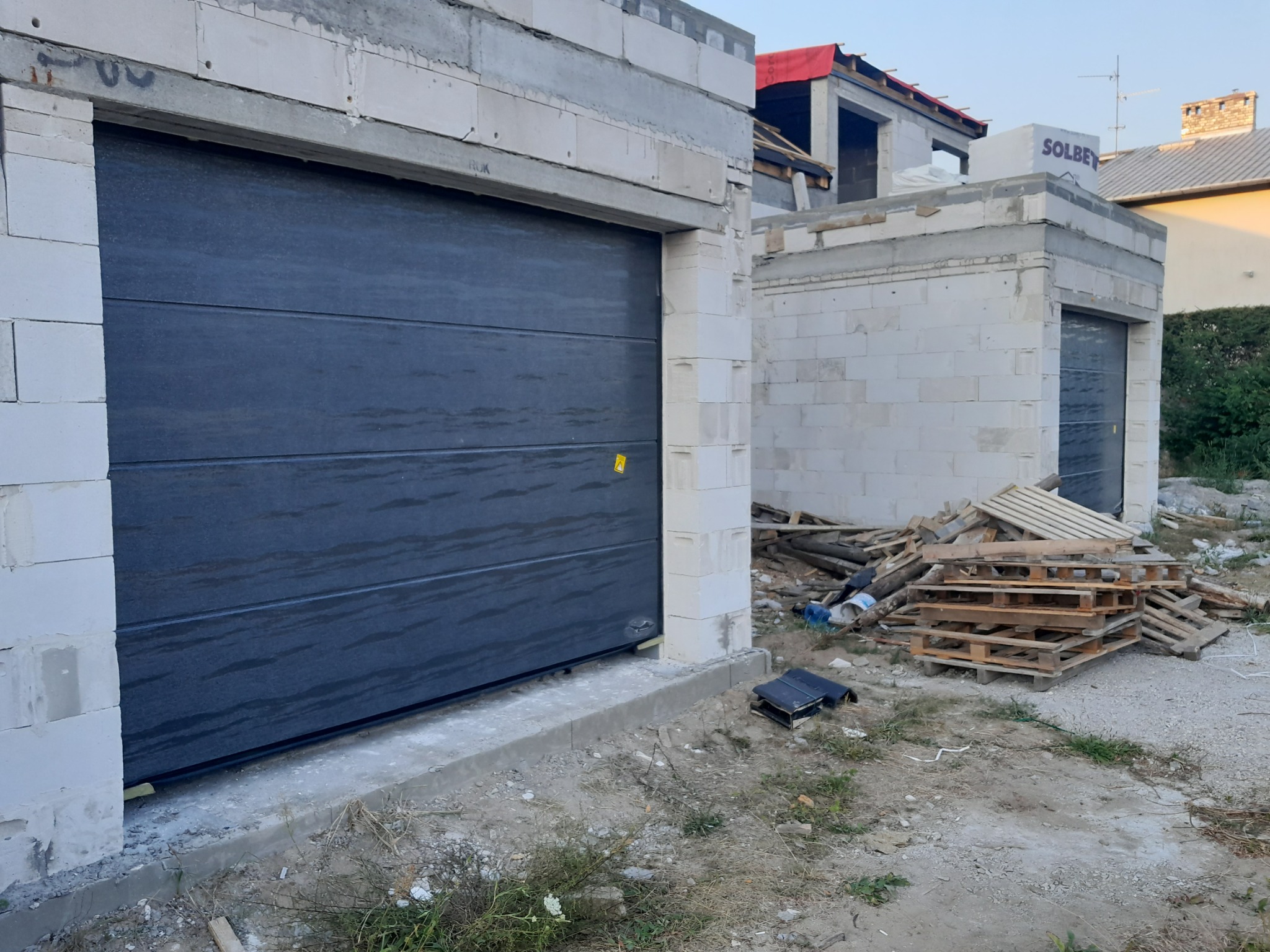 Szara segmentowa brama w surowym stanie, z widocznymi bloczkami betonowymi i stertą palet w tle, na placu budowy.