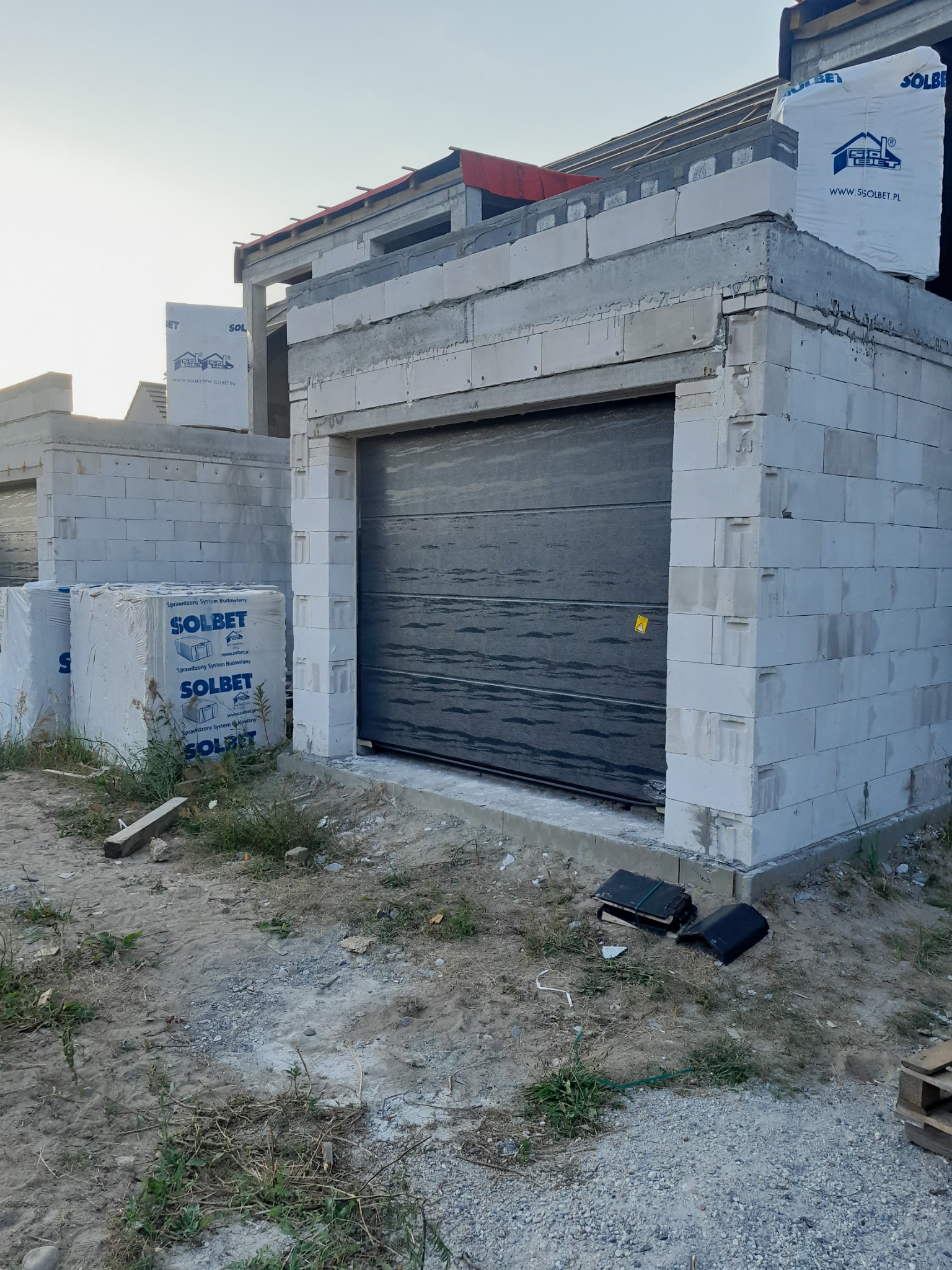 Nowa brama garażowa segmentowa w surowym stanie, zamontowana w budynku z bloczków Solbet w trakcie budowy, widoczne materiały budowlane i narzędzia.