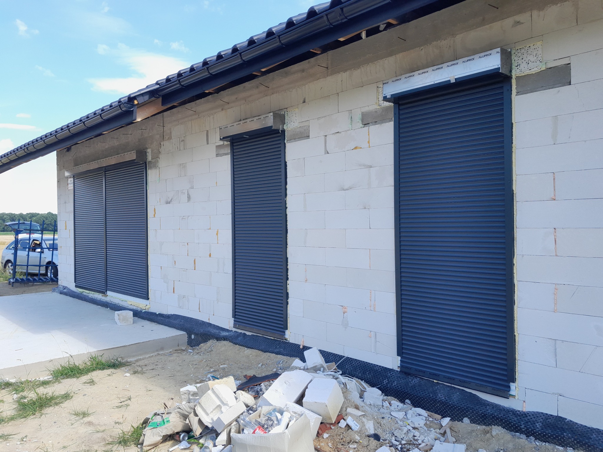 Nowo wybudowany dom z trzema oknami zabezpieczonymi szarymi roletami zewnętrznymi, widoczne materiały budowlane i samochód w tle.