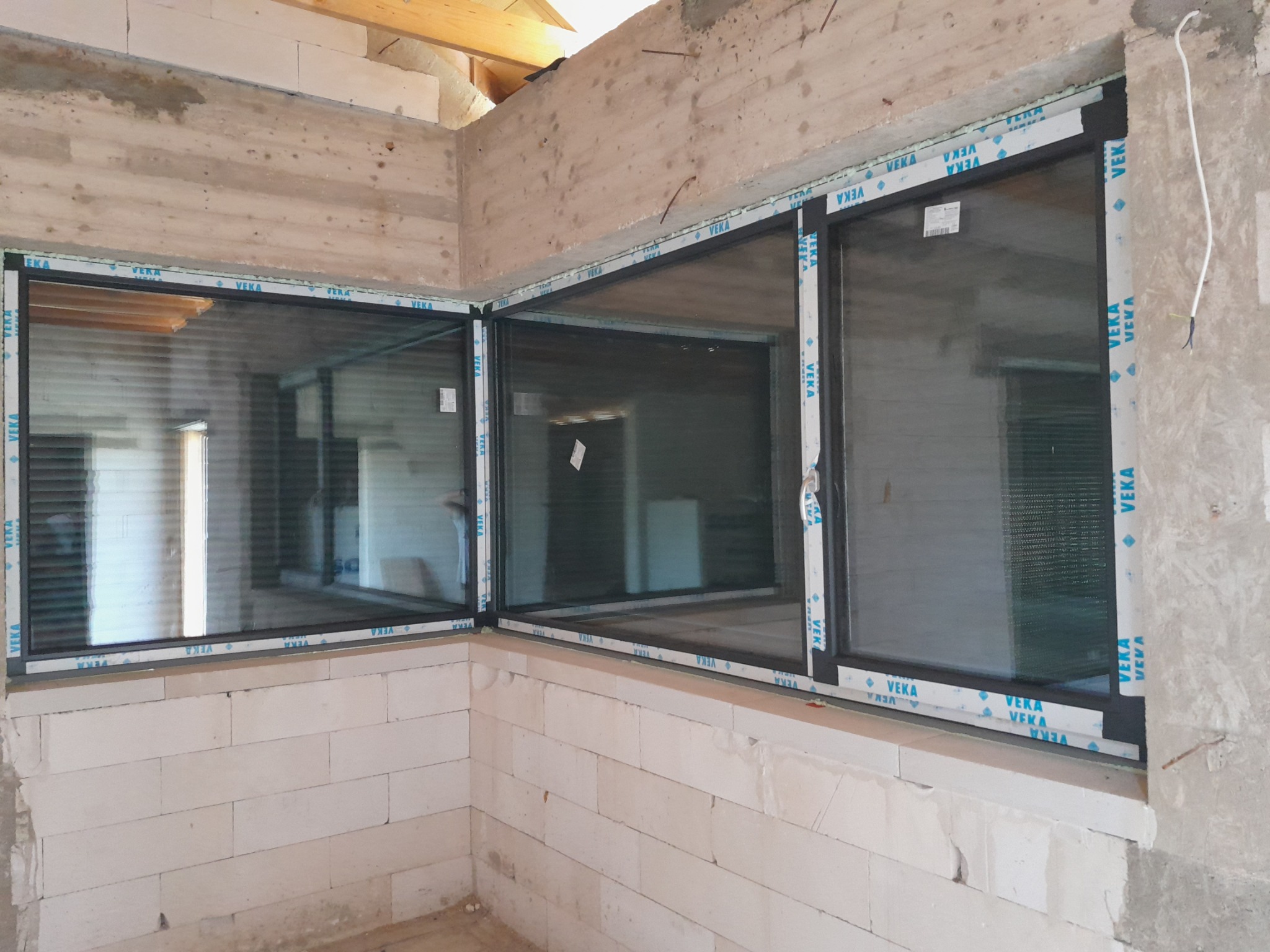 Montaż okien PCV w surowym stanie w Henrykowie. Widoczne trzy okna z czarnymi ramami, uszczelnione pianką montażową, z taśmą ochronną producenta na obrzeżach, zamontowane w ścianie z białych bloczków.