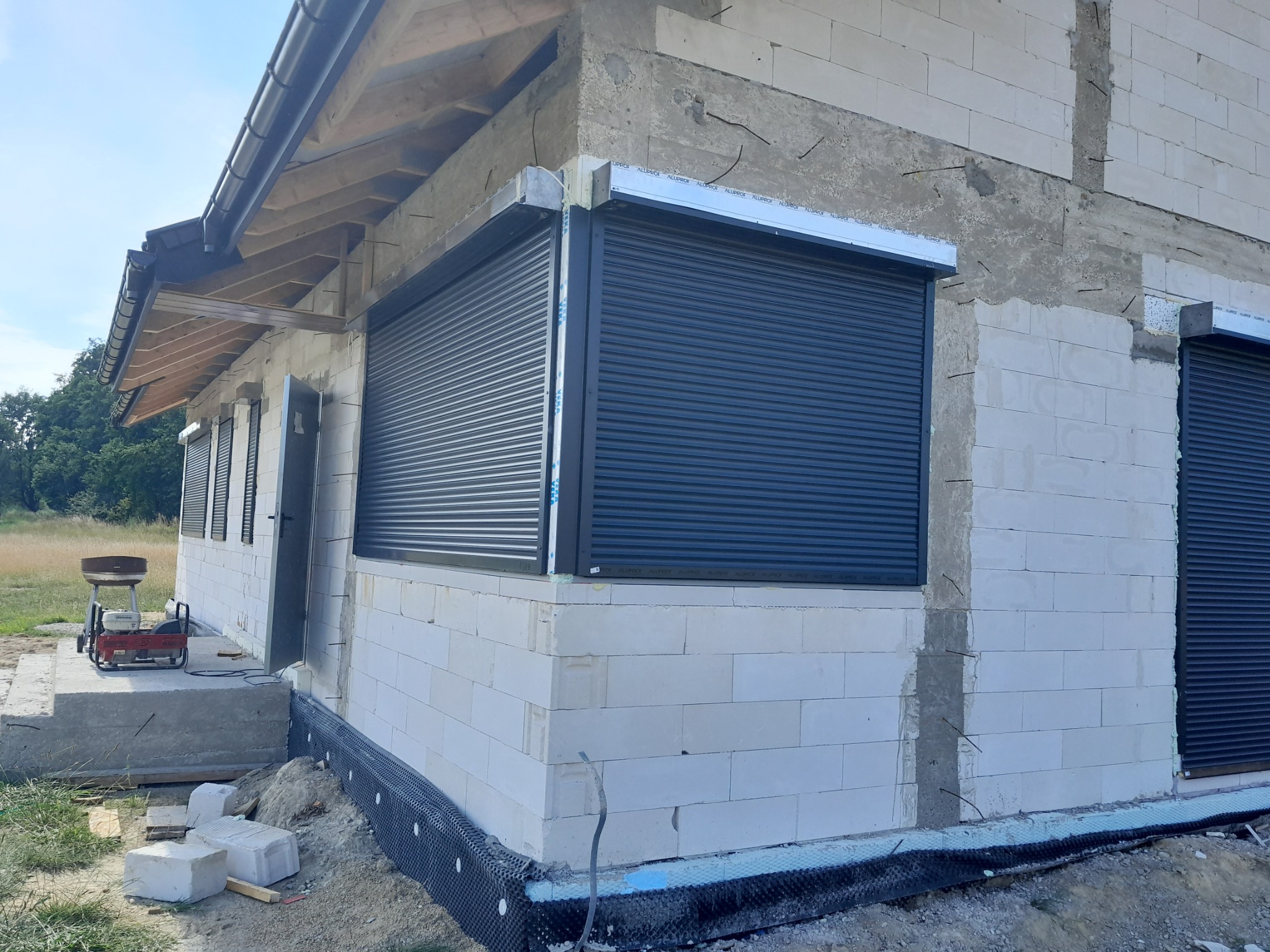 Budynek w trakcie budowy z zamontowanymi roletami zewnętrznymi w kolorze grafitowym, widoczne bloki betonowe, ocieplenie fundamentów i wystające pręty zbrojeniowe.