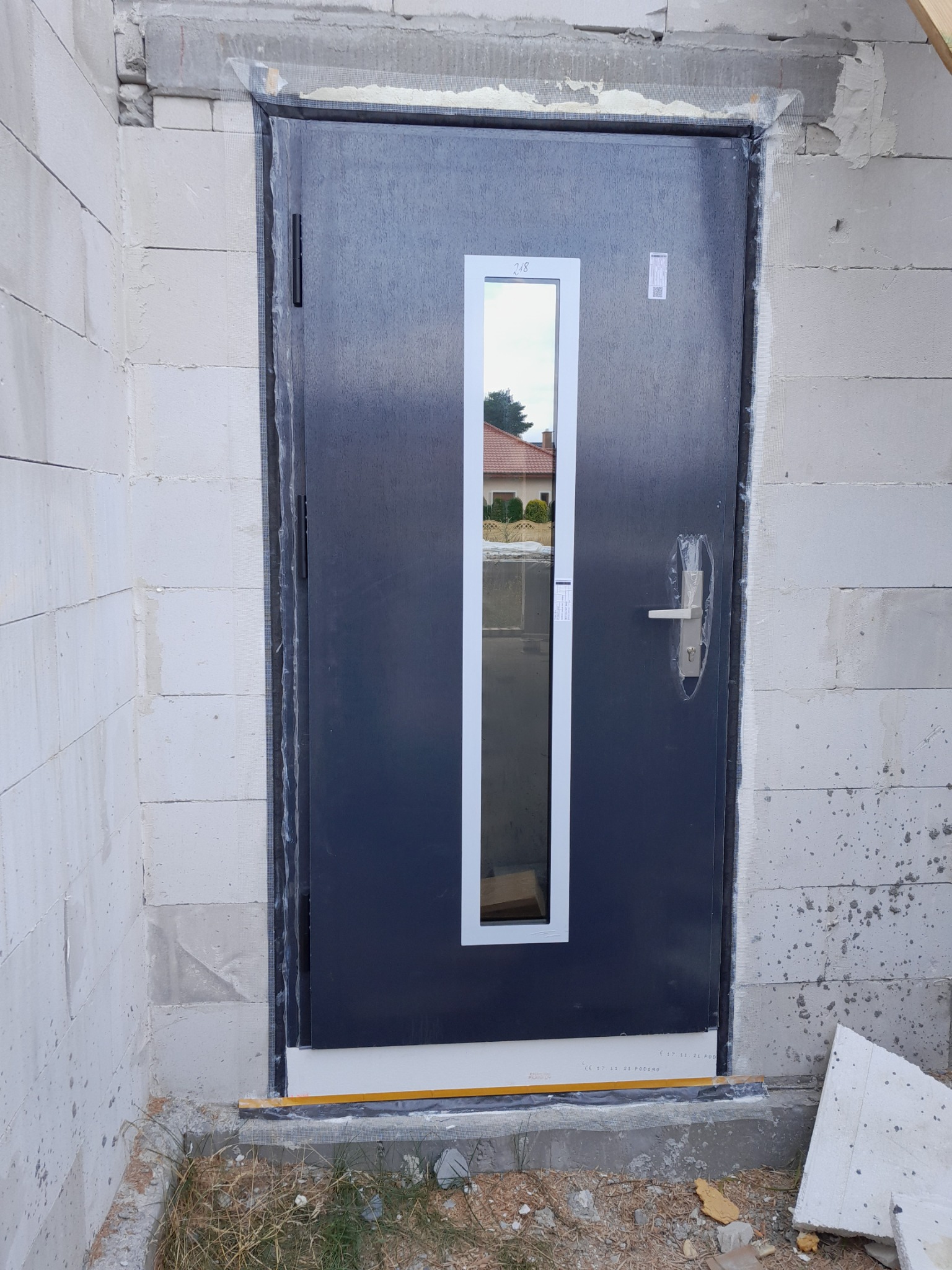 Ciemne drzwi zewnętrzne z wąskim przeszkleniem w białej ramie, zamontowane w surowym murze z betonu komórkowego, widoczne zabezpieczenia folią i taśmą montażową.