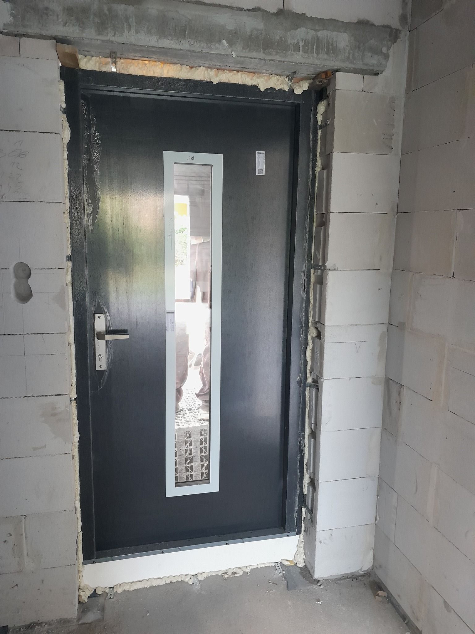 Ciemne drzwi wejściowe z długim, pionowym przeszkleniem zamontowane w surowym murze z białych bloczków, widoczna piana montażowa wokół ościeżnicy.