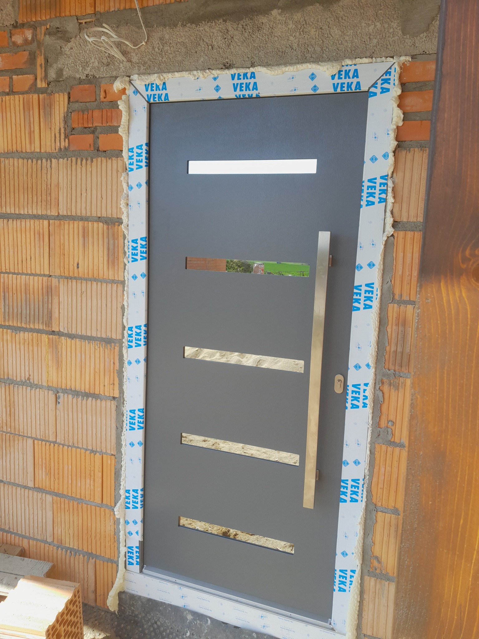 Szara, nowoczesna konstrukcja z długim, pionowym uchwytem, osadzona w surowej, ceglanej ścianie, z widoczną pianką montażową i folią ochronną na ramie.