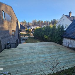 Patryk Marek Góral dom - Widok z góry na nowo wybudowany taras drewniany z desek w odcieniach zieleni i szarości, z widocznymi domami w tle i fragmentem skalnego podłoża w dolnej części kadru.