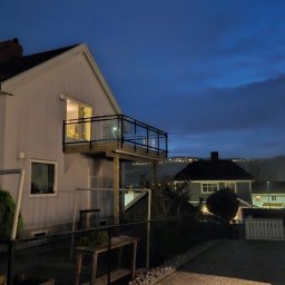 Patryk Marek Góral dom - Dom z drewnianym tarasem z czarną balustradą i szklanym wypełnieniem, widok na oświetlone miasto w oddali pod wieczornym niebem.