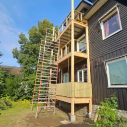 Wielopoziomowa konstrukcja tarasu drewnianego w trakcie budowy, z widocznymi rusztowaniami, surowym drewnem i elementami mocującymi, przy ciemnej elewacji domu z oknami.