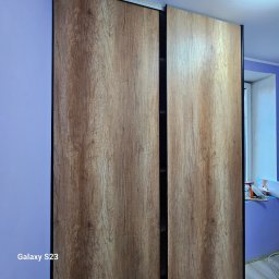 Patryk Marek Góral dom - Nowoczesna szafa przesuwna z drewnopodobnymi frontami w odcieniach brązu i czarnymi obramowaniami, zrealizowana w Siemianowicach Śląskich. Widoczny fragment ściany pomalowanej na fioletowo i okno...