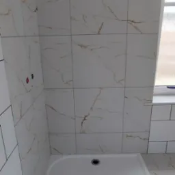 Nowa kabina prysznicowa z białym brodzikiem i płytkami imitującymi marmur na ścianach, widoczne fragmenty instalacji wodnej.